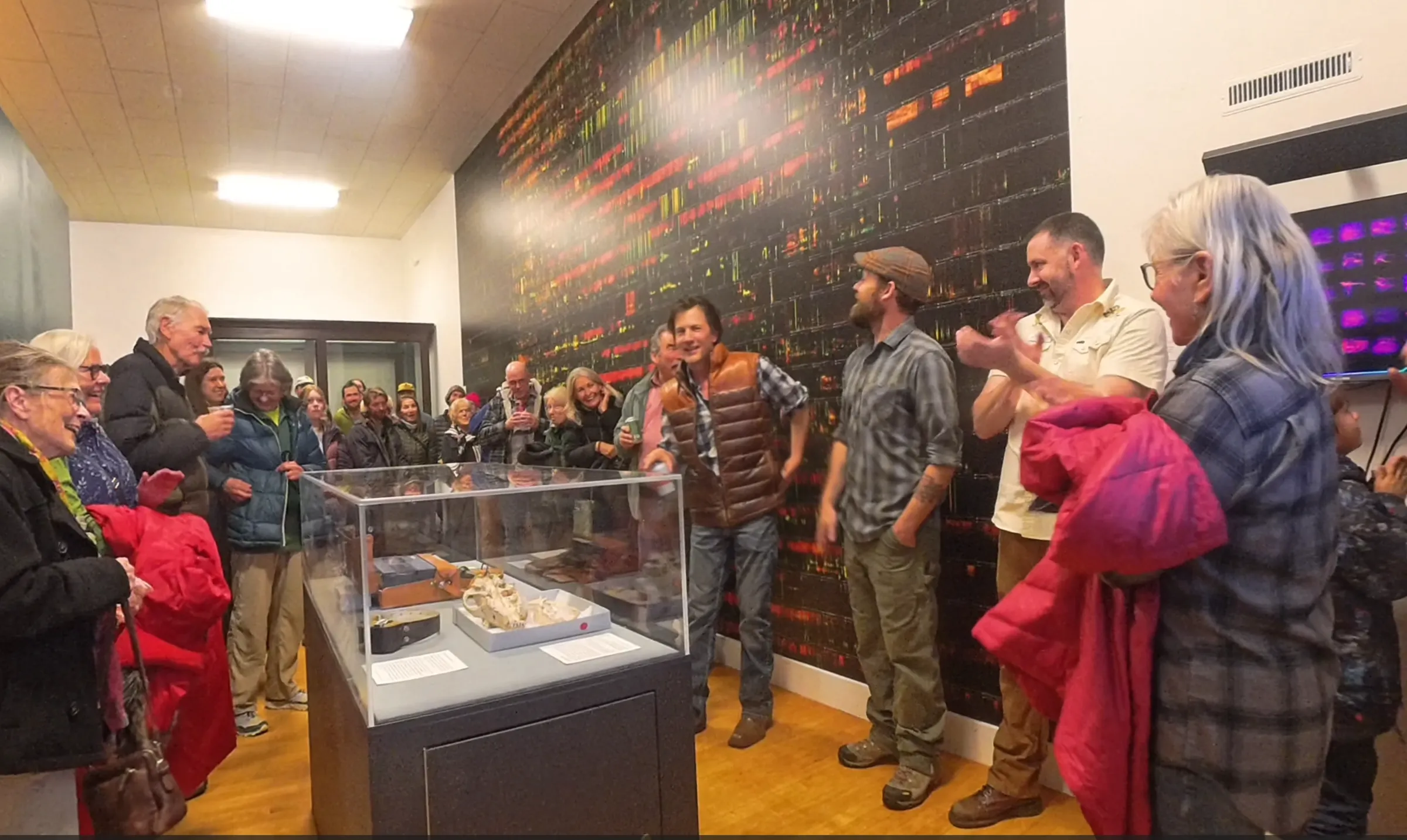 A crowd gathers in a gallery beside a glass display case holding a wolf skull and a collar, in front of a wall-sized dark spectrogram streaked with red and yellow.