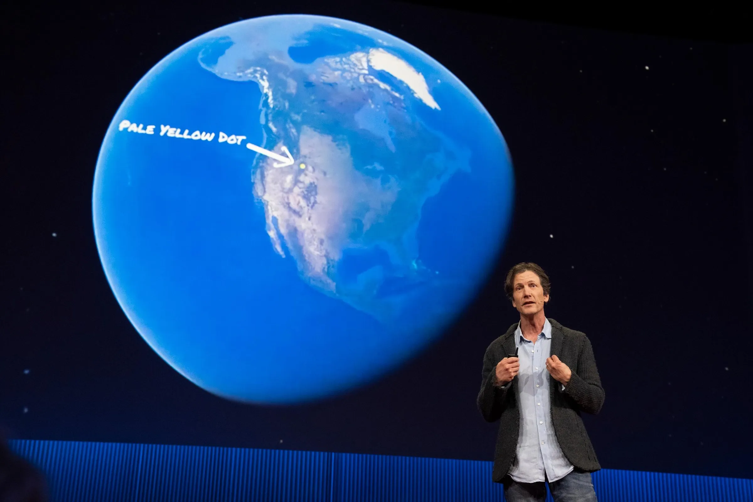 Jeff Reed speaks on a dark stage in front of a large screen showing North America from space, with an arrow labeled 'Pale yellow dot' pointing to a spot in the northern Rockies.