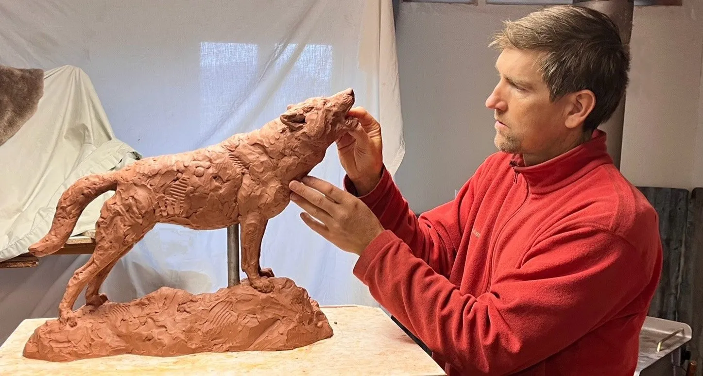 A man in a red fleece shapes the head of a clay sculpture of a howling wolf standing on a rocky base.