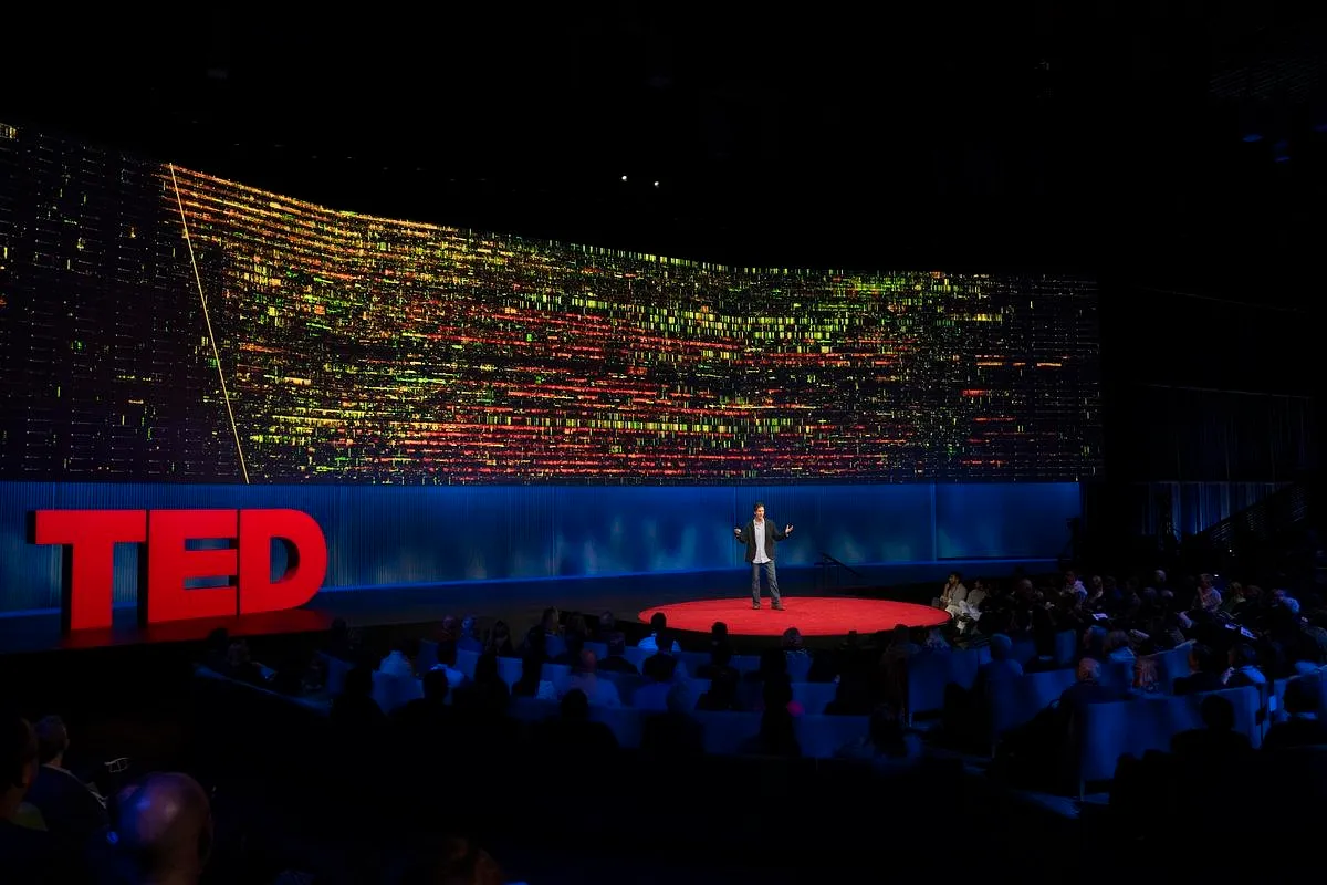 Jeff Reed on the TED stage, with a wall-size spectrogram glowing yellow and red across the screen behind him.