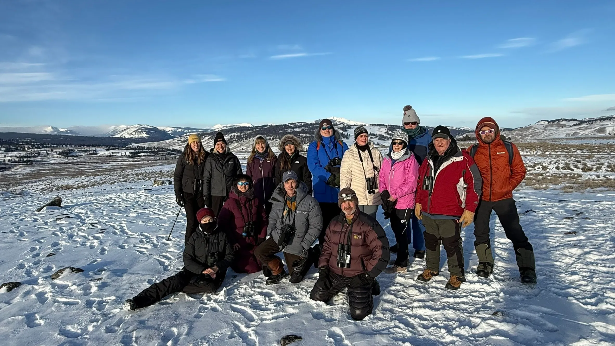 A group of about fifteen people in winter jackets, many with binoculars, pose on a snowy rise with mountains and blue sky behind them.
